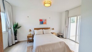Een slaapkamer met een groot bed, twee nachtkastjes, een bijzettafeltje, een potplant, een kunstwerk aan de muur en een glazen schuifdeur naar een buitenruimte.