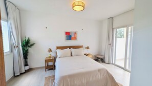 A bedroom with a large bed, two bedside tables, a nightstand, a potted plant, a wall art piece, and a sliding glass door leading to an outdoor area. - Dominicus Beach Haven (Dominicus)