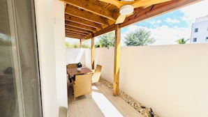 Een overdekt terras buiten, met een tafel en stoelen, houten balken en een witte muur.