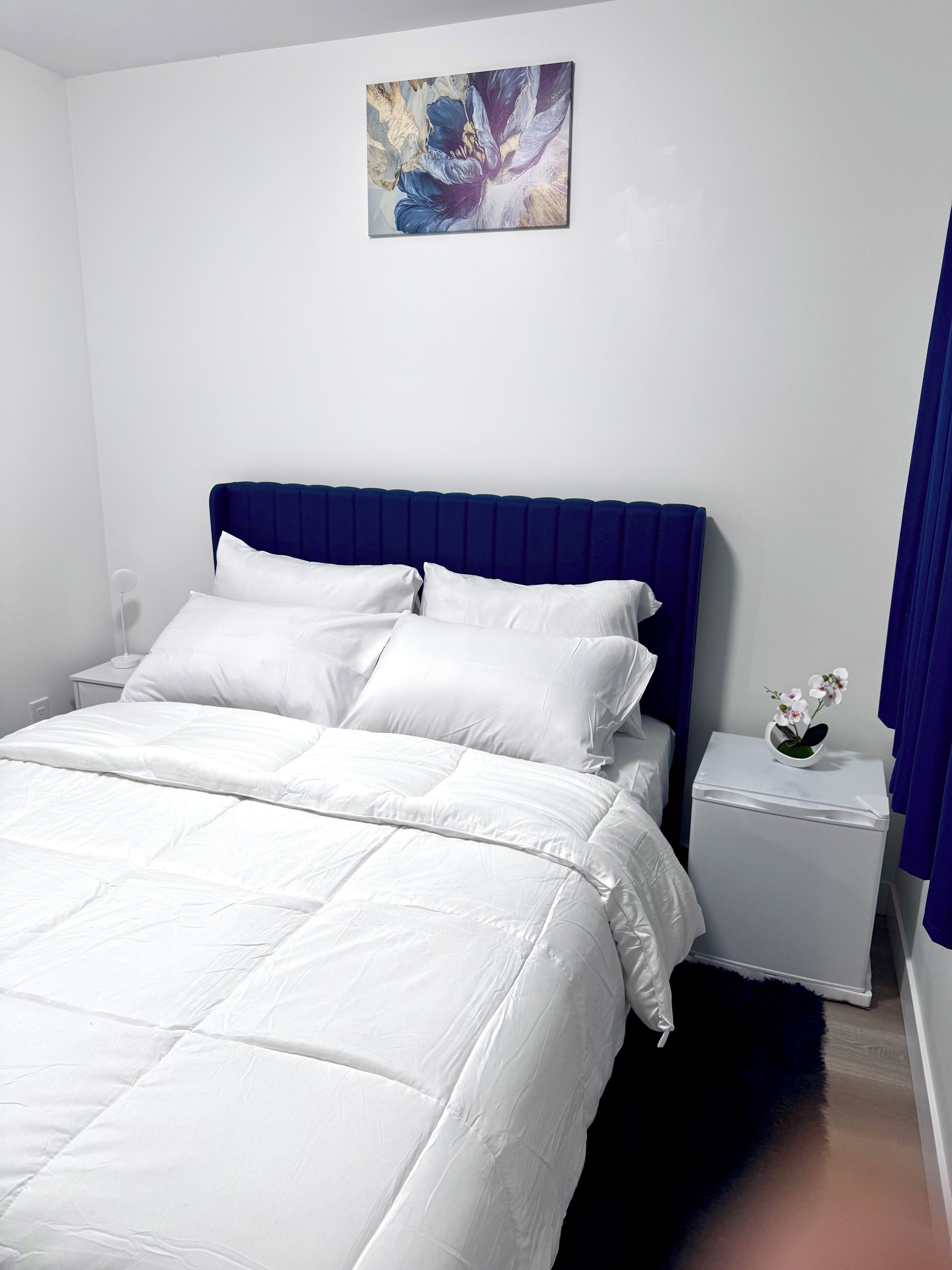 Een slaapkamer met een bed, witte beddengoed, een blauw hoofdeinde, een nachtkastje met een witte vaas en een schilderij aan de muur.