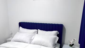 Een slaapkamer met een bed, witte beddengoed, een blauw hoofdeinde, een nachtkastje met een witte vaas en een schilderij aan de muur.