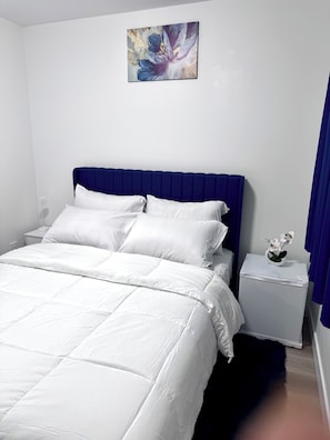 Une chambre à coucher avec un lit, du linge de lit blanc, une tête de lit bleue, une table de chevet avec un vase blanc et un tableau accroché au mur.