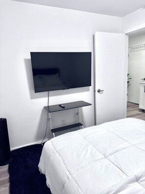 A bedroom with a bed, a TV mounted on the wall, a door, and a small shelf. - Cozy Central Winnipeg Main Floor Suite – Private & Quiet (Winnipeg)