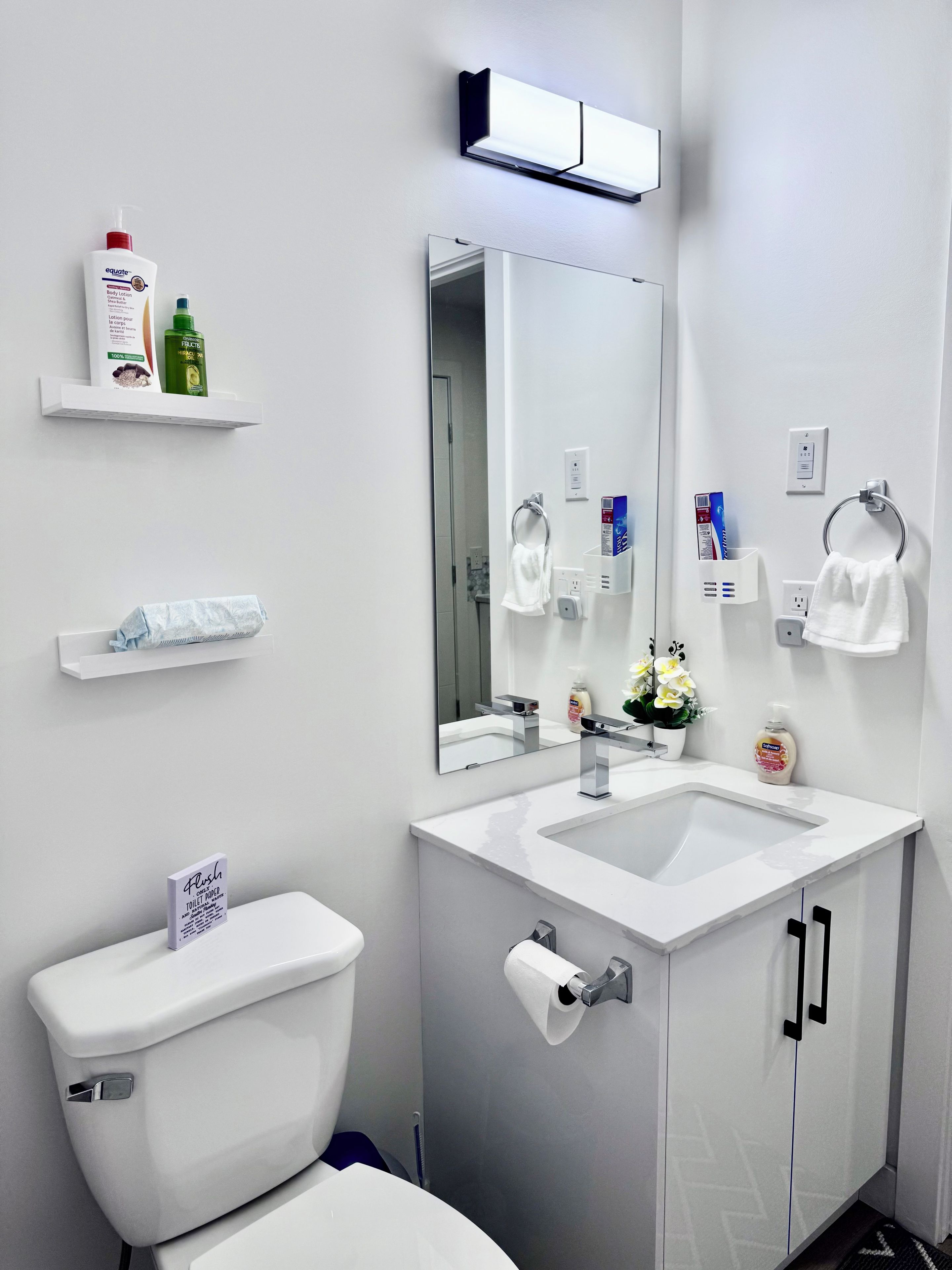 Une salle de bain moderne avec un lavabo blanc, un miroir et des toilettes. On y trouve des étagères avec des articles de toilette et un porte-serviettes.