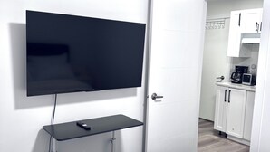 Une chambre d’hôtel avec un téléviseur à écran plat fixé au mur, un lit avec une literie blanche et, au fond, une petite cuisine.
