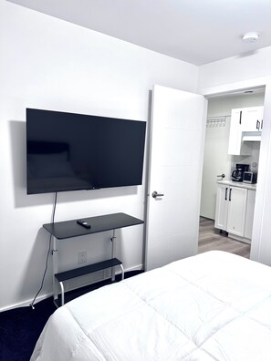 A hotel room with a flat-screen TV mounted on the wall, a bed with white bedding, and a kitchenette in the background. - Cozy Central Winnipeg Main Floor Suite – Private & Quiet (Winnipeg)