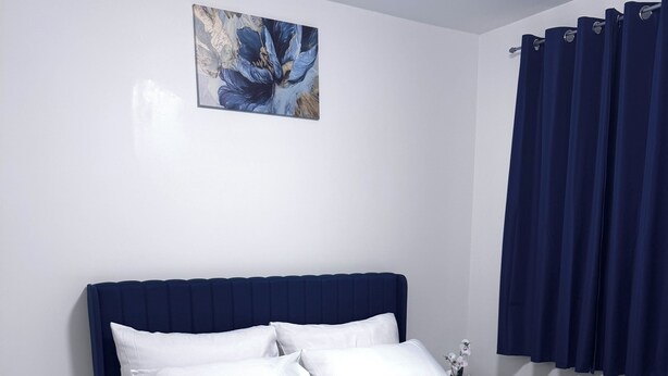 Un lit bien fait, avec une literie blanche, une tête de lit bleue et une table de chevet sur laquelle se trouve une horloge.