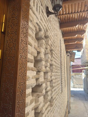A corridor with a textured brick wall, a wooden door with intricate carvings, and a hanging light fixture. - ANIS-PARI Hotel (Bukhara)