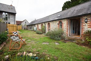 Un jardin avec une pelouse, une chaise en bois, un mur de pierres et un bâtiment en briques doté d’une porte.