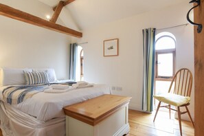 Ein Schlafzimmer mit einem Bett, einer Holzbank, einem Stuhl, einem Fenster mit Vorhängen und einem Bild an der Wand.
