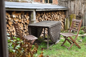Ein Außenbereich mit Sitzgelegenheit, einem Tisch, zwei Stühlen, einem Holzstapel und einer rustikalen Holzhütte im Hintergrund.
