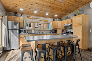 A modern kitchen with wooden cabinets, a stainless steel refrigerator, a large island with bar stools, and a wooden ceiling.