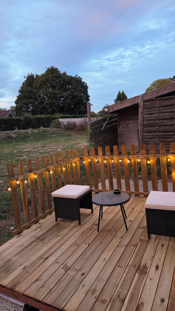 A wooden deck with two wicker chairs and a small table, surrounded by a wooden fence with string lights.