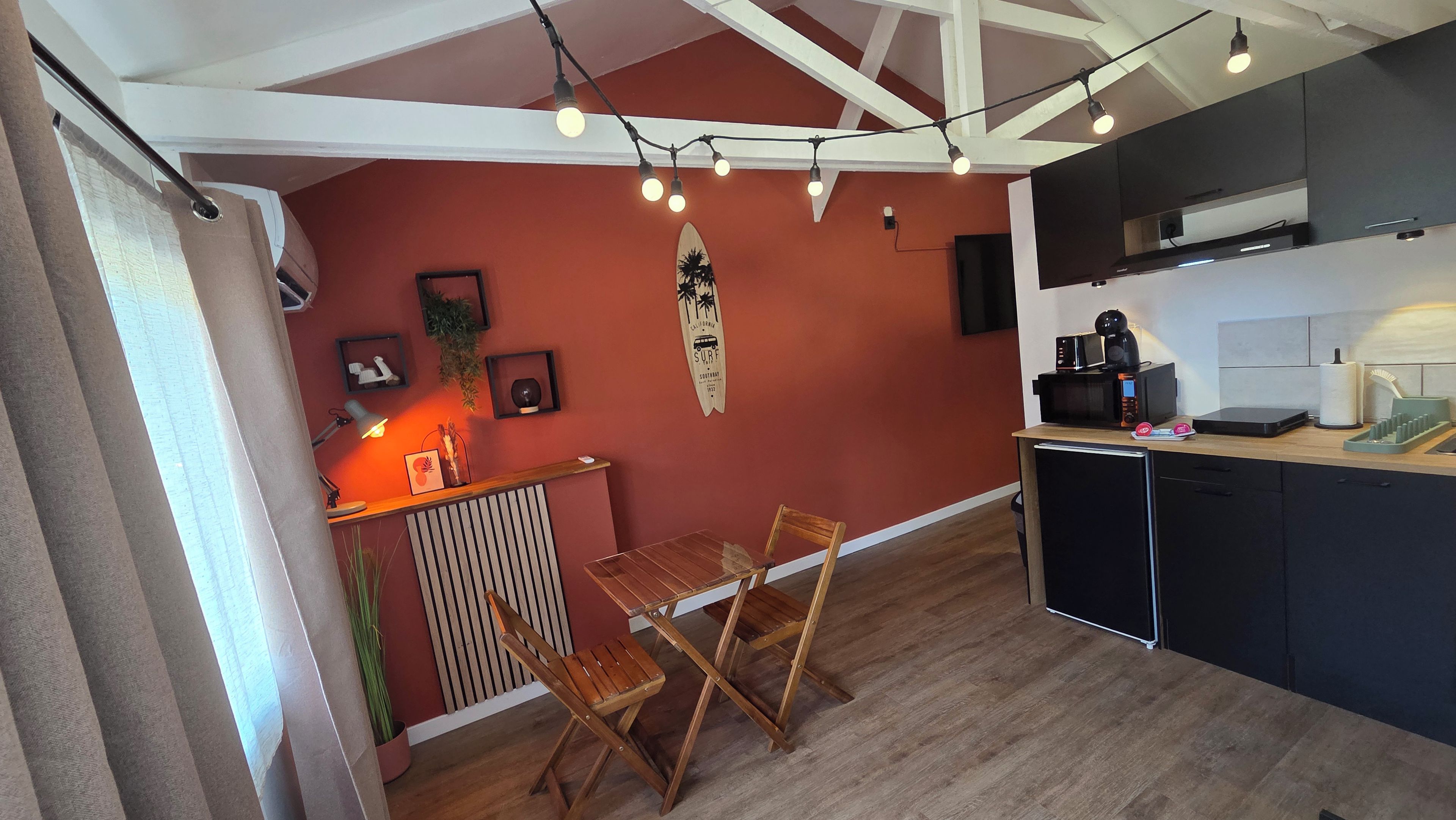 A compact kitchen with a dining area, featuring a wooden table and chairs, a surfboard decoration, and modern appliances.