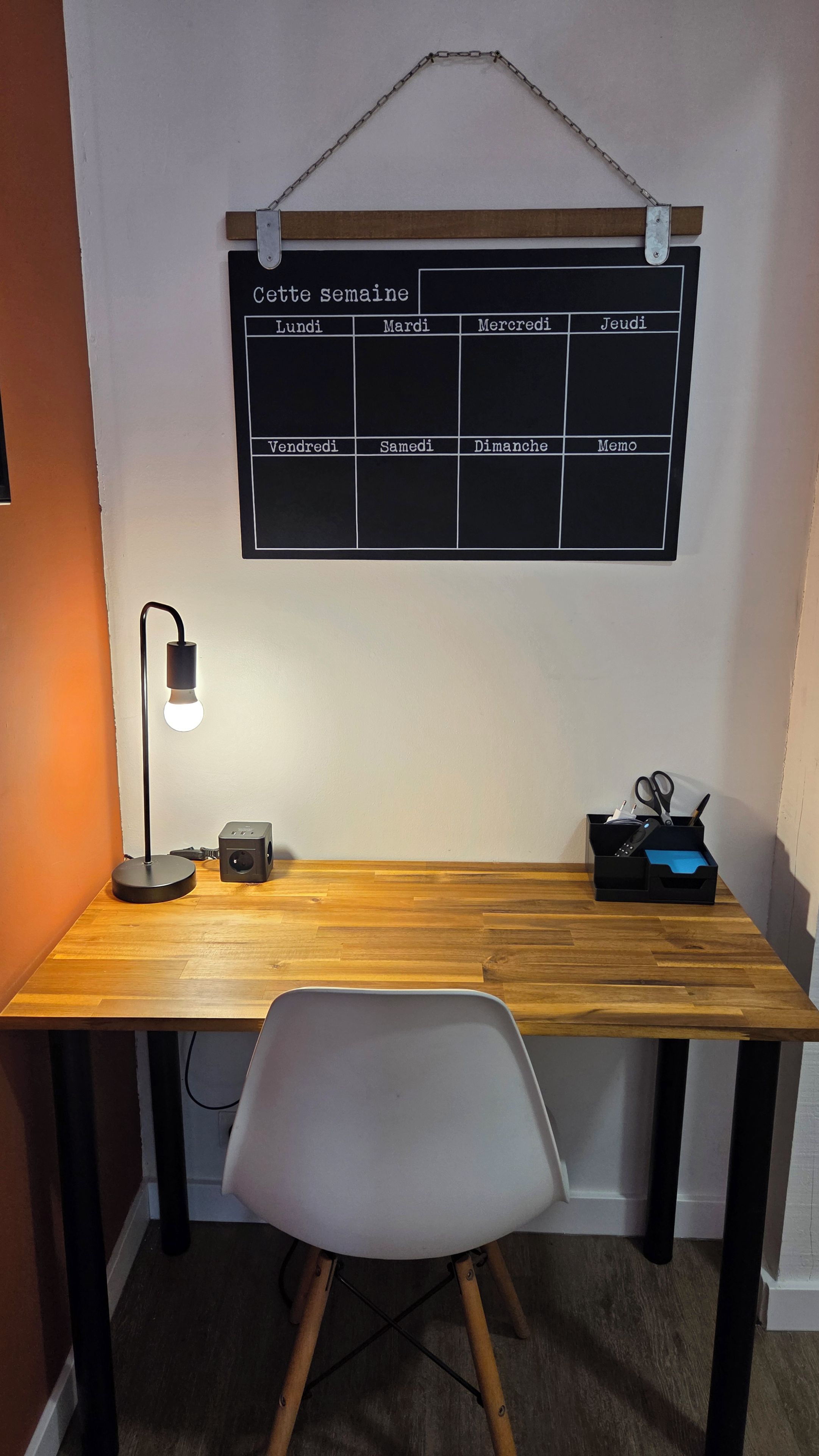 Een houten bureau met een witte stoel, een bureaulamp en een zwarte speaker. Daarboven hangt een schoolbord waarop de dagen van de week in het Italiaans staan.