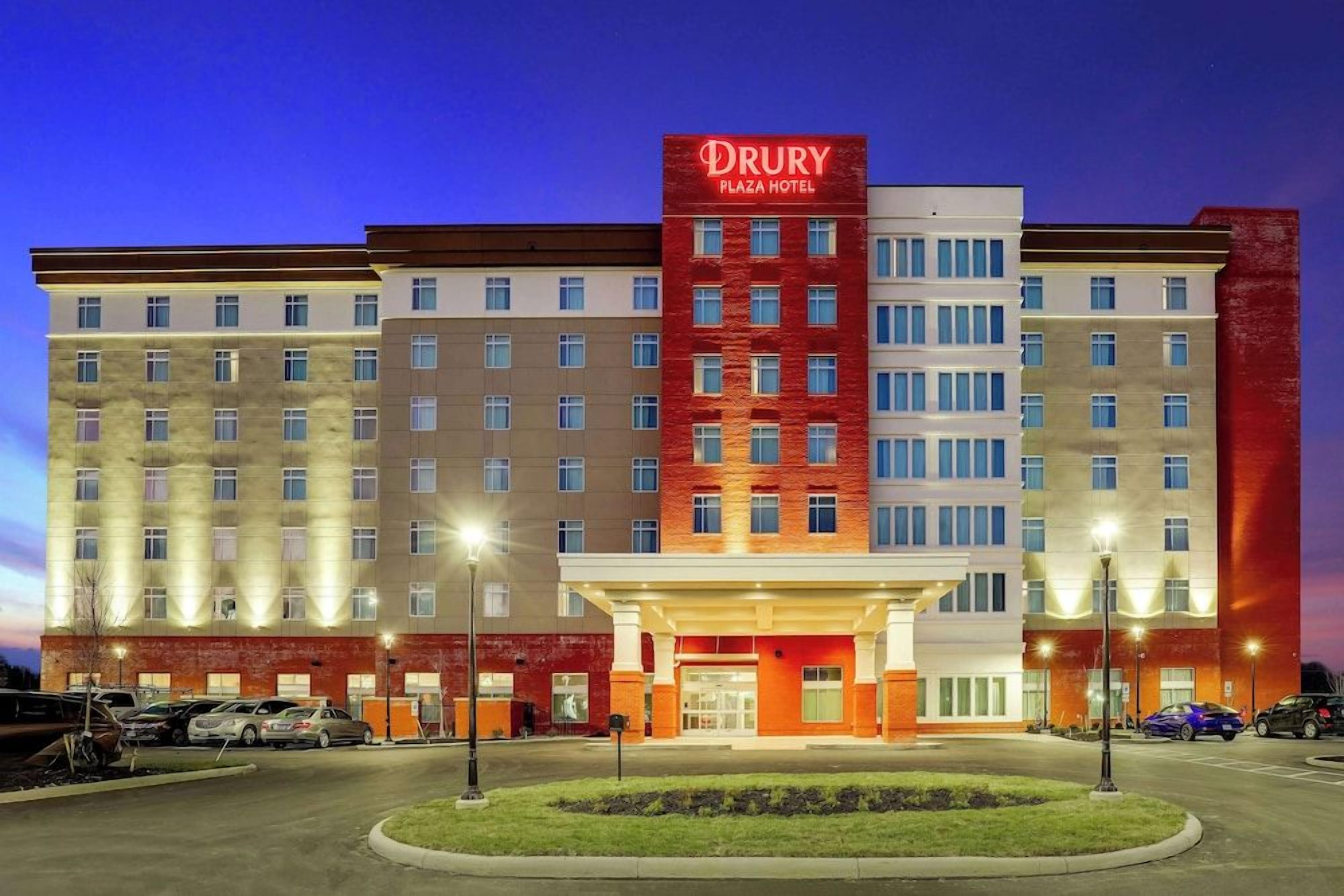 A Drury Plaza Hotel with a prominent red and white facade, multiple floors, and a parking lot with several cars.