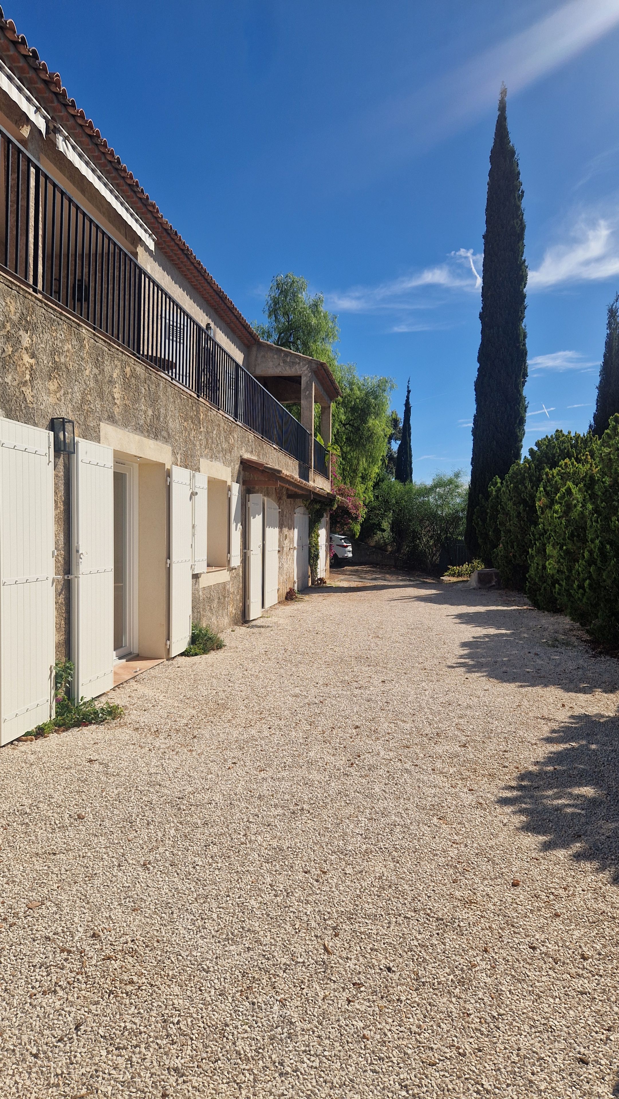 Un bâtiment avec une allée gravillonnée, un balcon doté d’une balustrade et un grand arbre en arrière-plan.