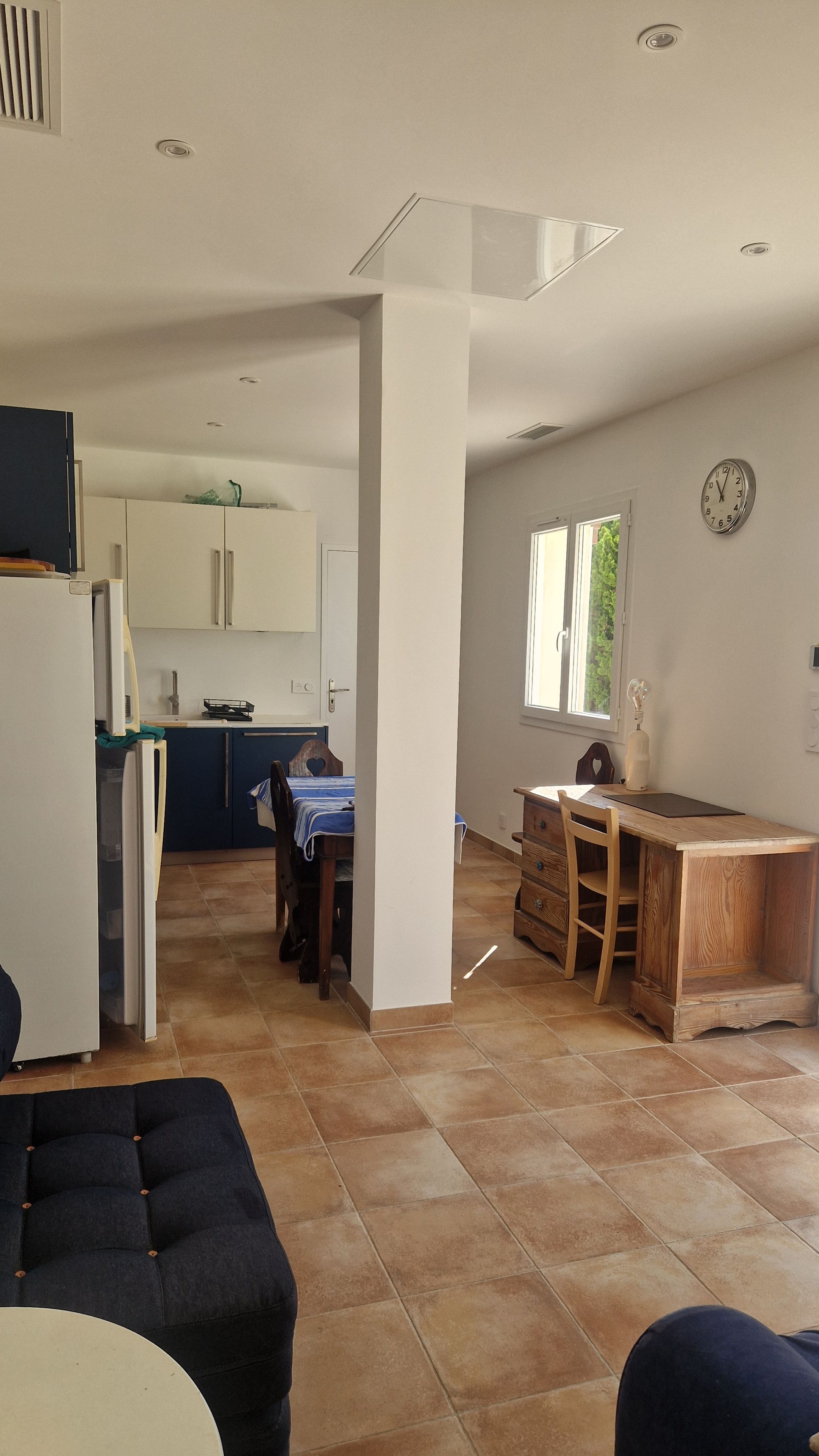 A spacious living area with a dining table, a kitchen in the background, and a large window providing natural light.