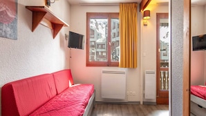 Una habitación compacta con un sofá rojo, una mesita, un televisor montado en la pared y una ventana con cortinas.