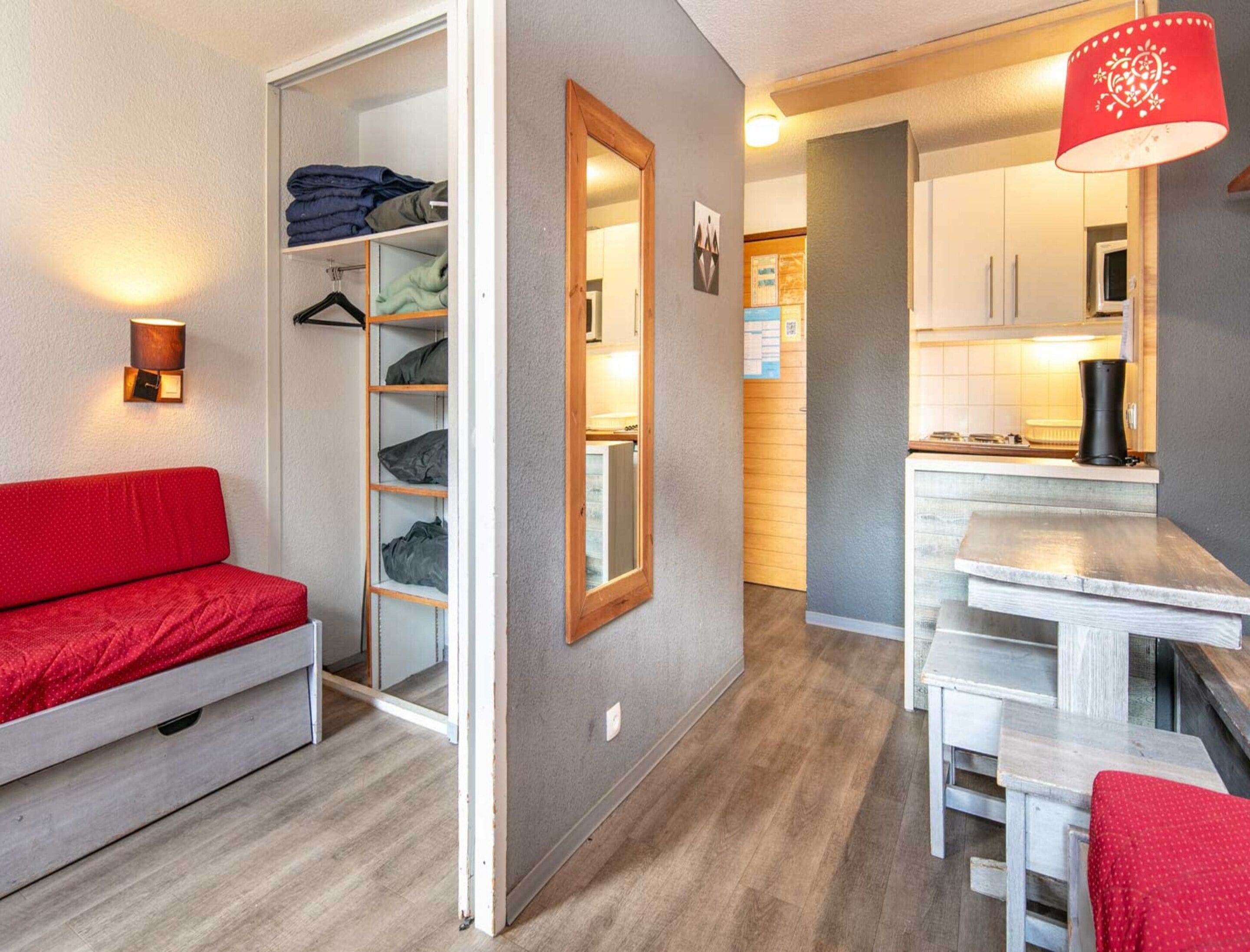 Een kleine leefruimte met een kitchenette, een rode bank, een houten laddenplank en een rode lamp.