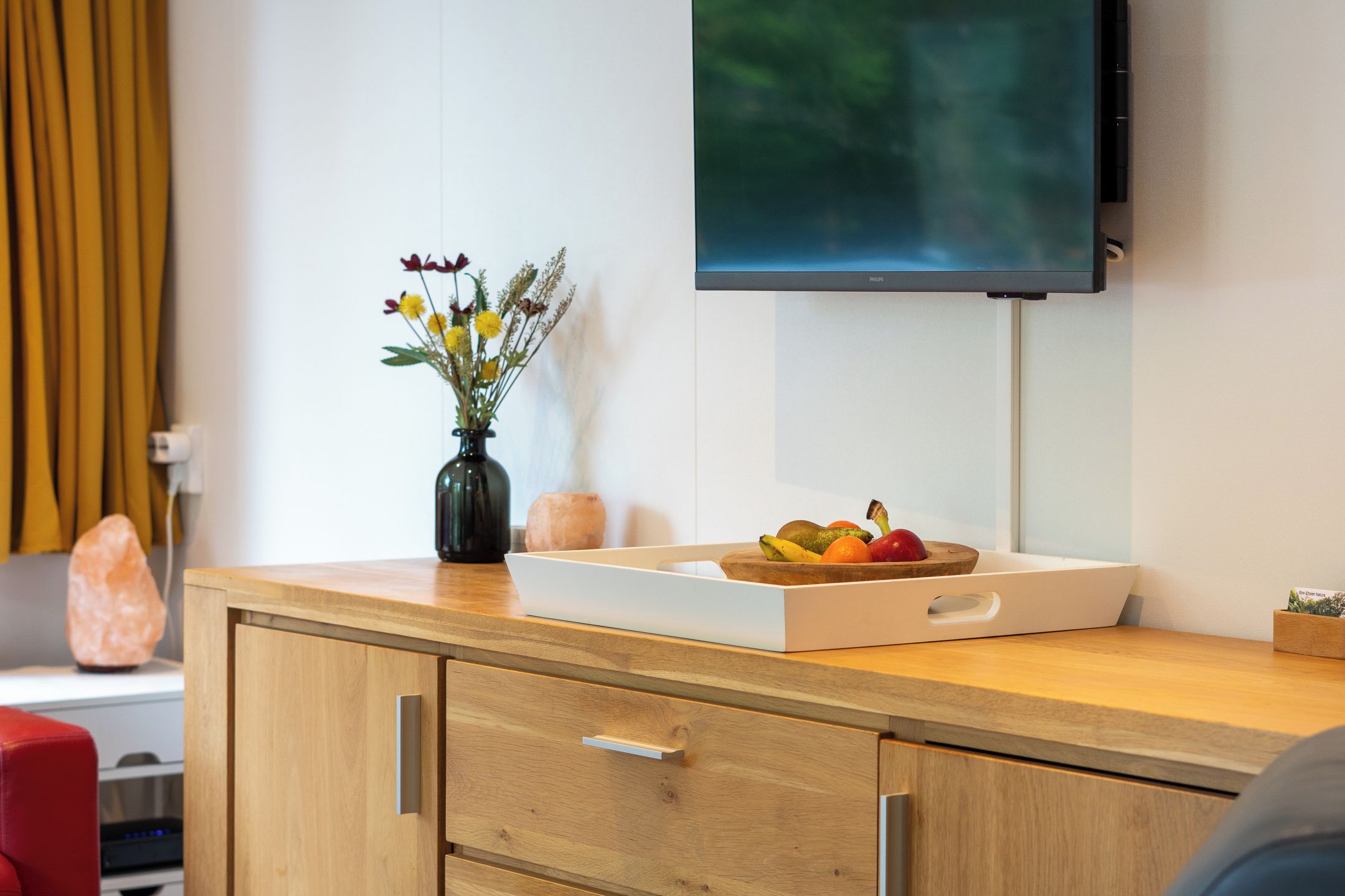 Un meuble en bois, avec un téléviseur à écran plat fixé au mur, un vase rempli de fleurs et un plateau de fruits posé sur le dessus du meuble.