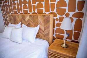 Ein Bett mit weißer Bettwäsche und Kissen, ein Holzkopfbrett mit Fischgrätmuster und ein Nachttisch mit einer Lampe.