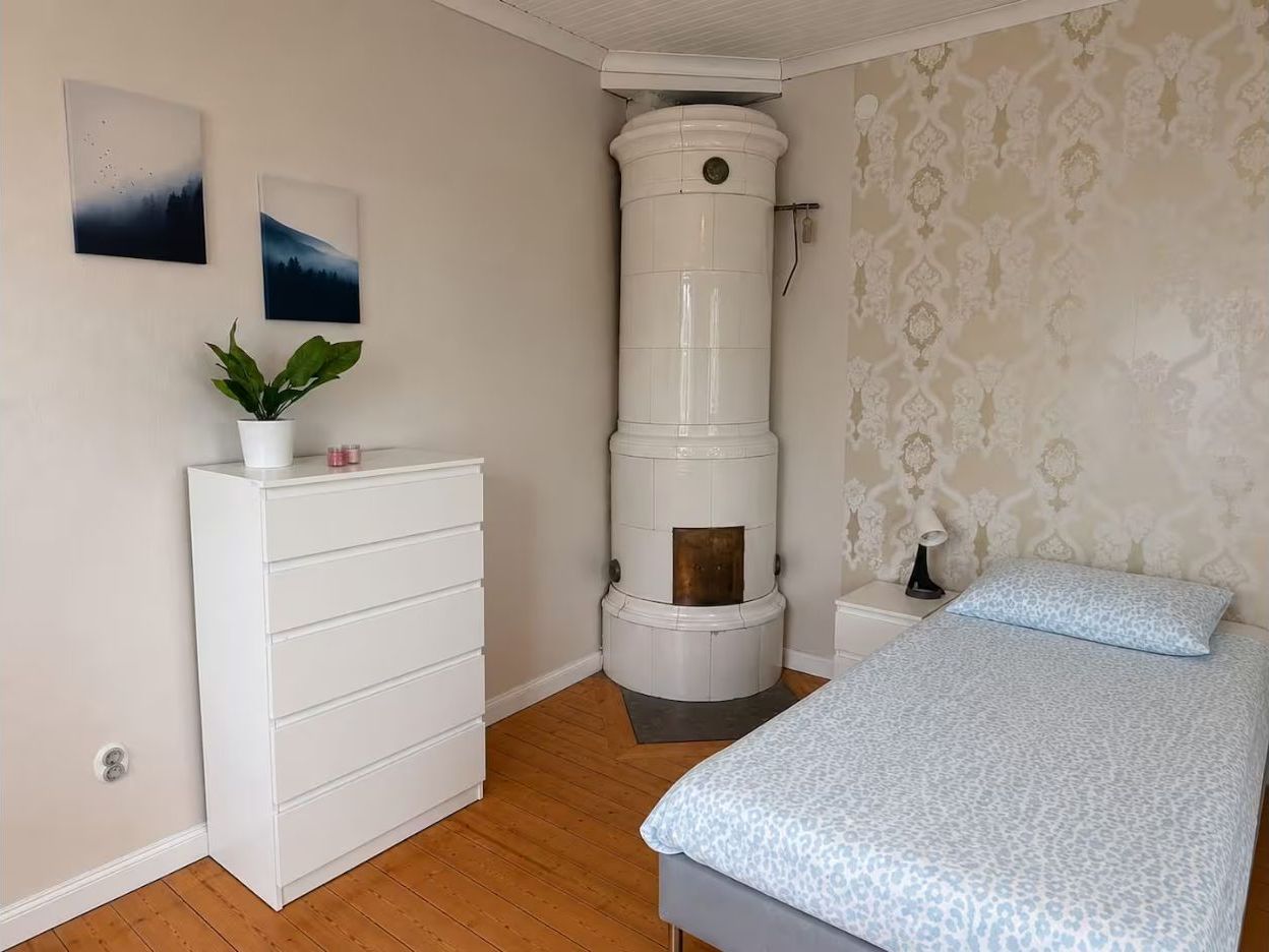 Ein Schlafzimmer mit einem Bett, einer Kommode, einer weißen Wand mit zwei Bildern, einem weißen Heizgerät und Wandpapier mit Muster.
