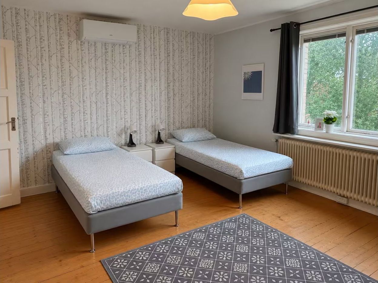 Ett sovrum med två sängar, ett element, ett fönster med gardiner och en mönstrad matta.