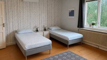 Ett sovrum med två sängar, ett element, ett fönster med gardiner och en mönstrad matta.