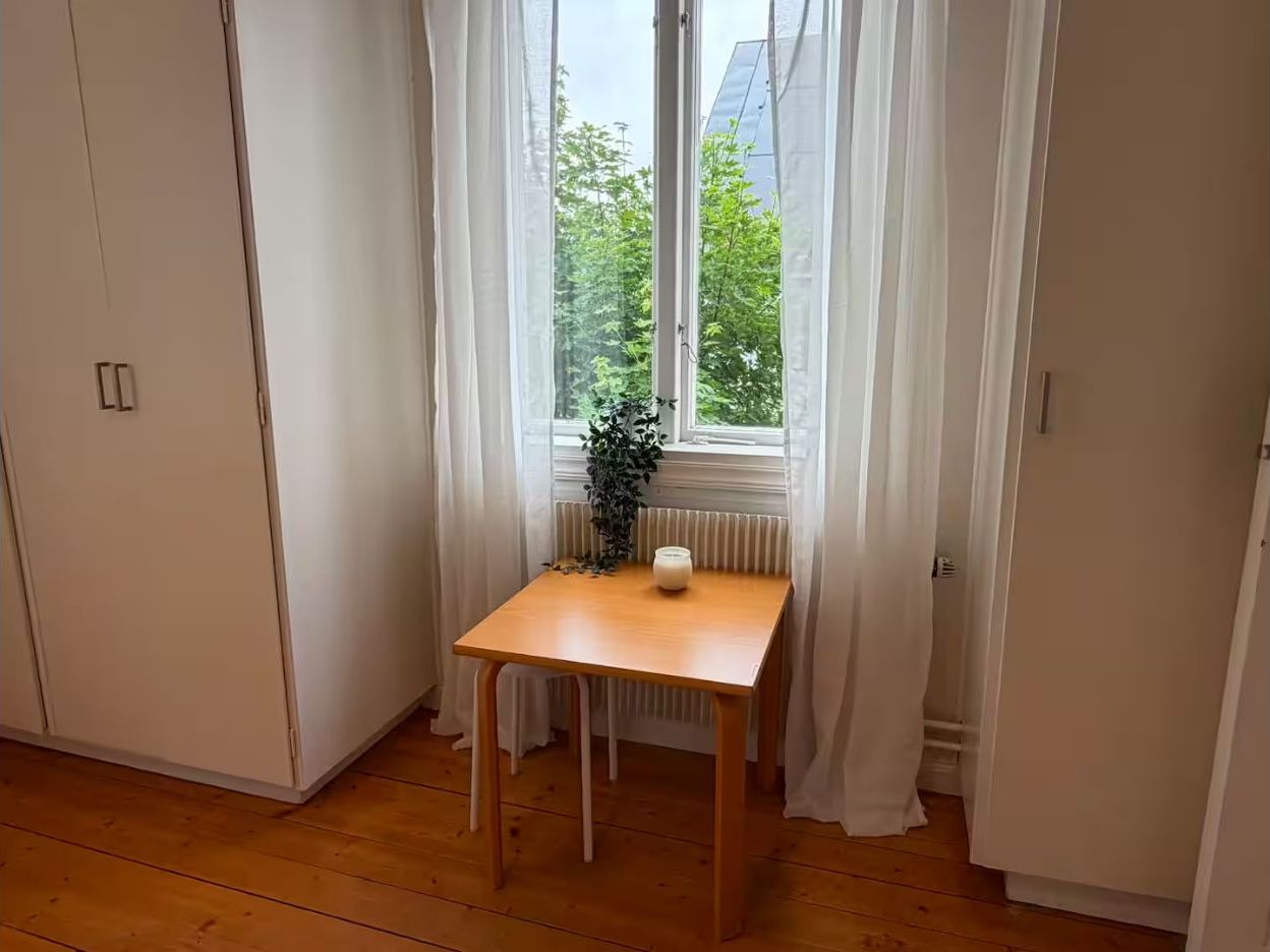 Huoneessa on puulattia, pöydällä palaa kynttilä ja ikkunassa on verhot.