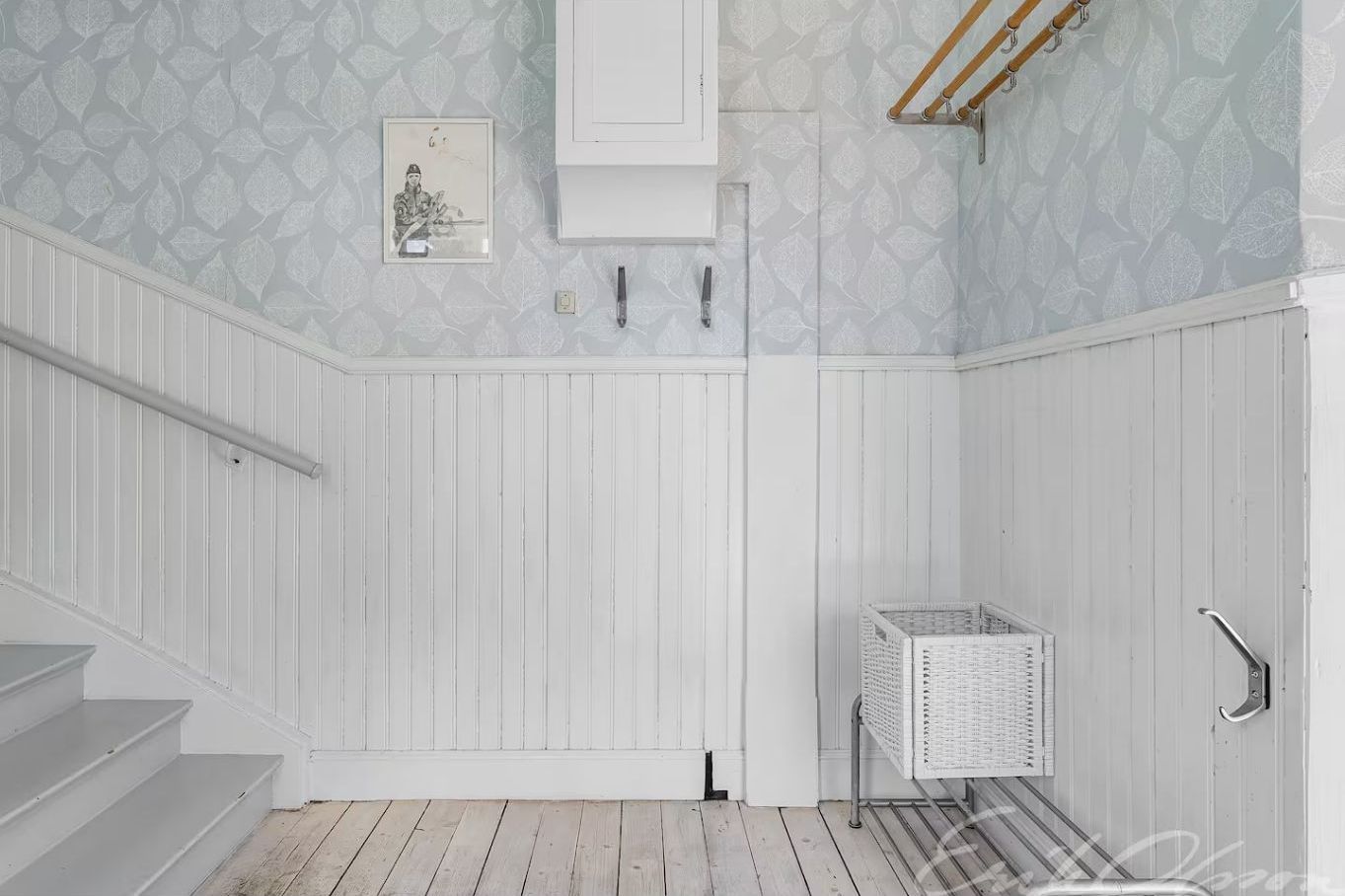 Ein Zimmer mit weißen Wandverkleidungen, einer Treppe, einem weißen Korb und einem gerahmten Bild an der Wand.