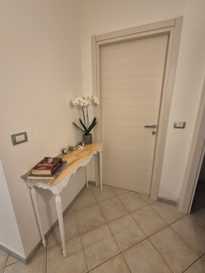 Un pasillo con una puerta blanca, una mesita con una planta y libros, y piso de baldosas.