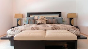 A bed with a patterned bedspread, two bedside lamps, and a bench at the foot of the bed. - HBK Villa Rentals (Jolly Harbour)