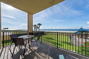 A balcony with a glass table and chairs, overlooking a beach with palm trees and a clear sky. - 🌊 Beachfront Gulf Views | Indoor & Outdoor Pools | Phoenix V 116 (Orange Beach)