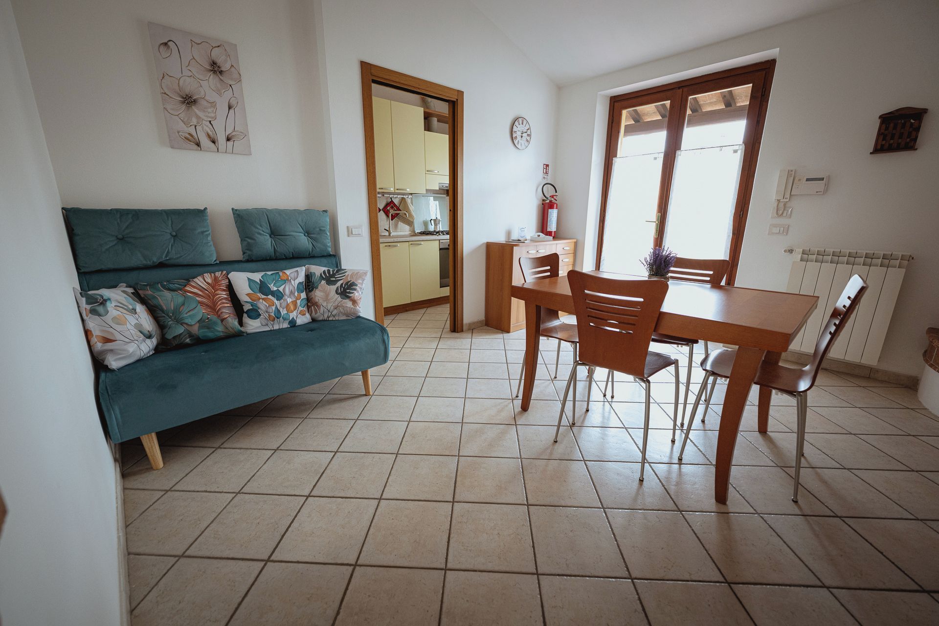 A living room with a teal sofa, a dining table with chairs, a kitchen area, and a large window.