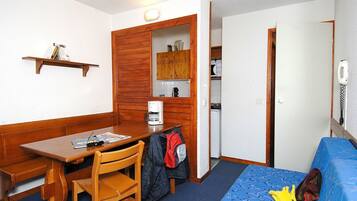 A compact room with a wooden table, a chair, a blue sofa, a small kitchenette, and a door leading to another room.