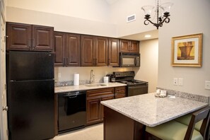 A kitchen with dark wooden cabinets, a black refrigerator, a microwave, and a granite countertop. - Chase Suite Hotel Lincoln (Lincoln)