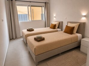 A hotel room with two beds, a window with curtains, a nightstand, and a wall-mounted light. - Spacious 2-Bedroom Apartment with Air Conditioning Near Laginha Beach (Mindelo)