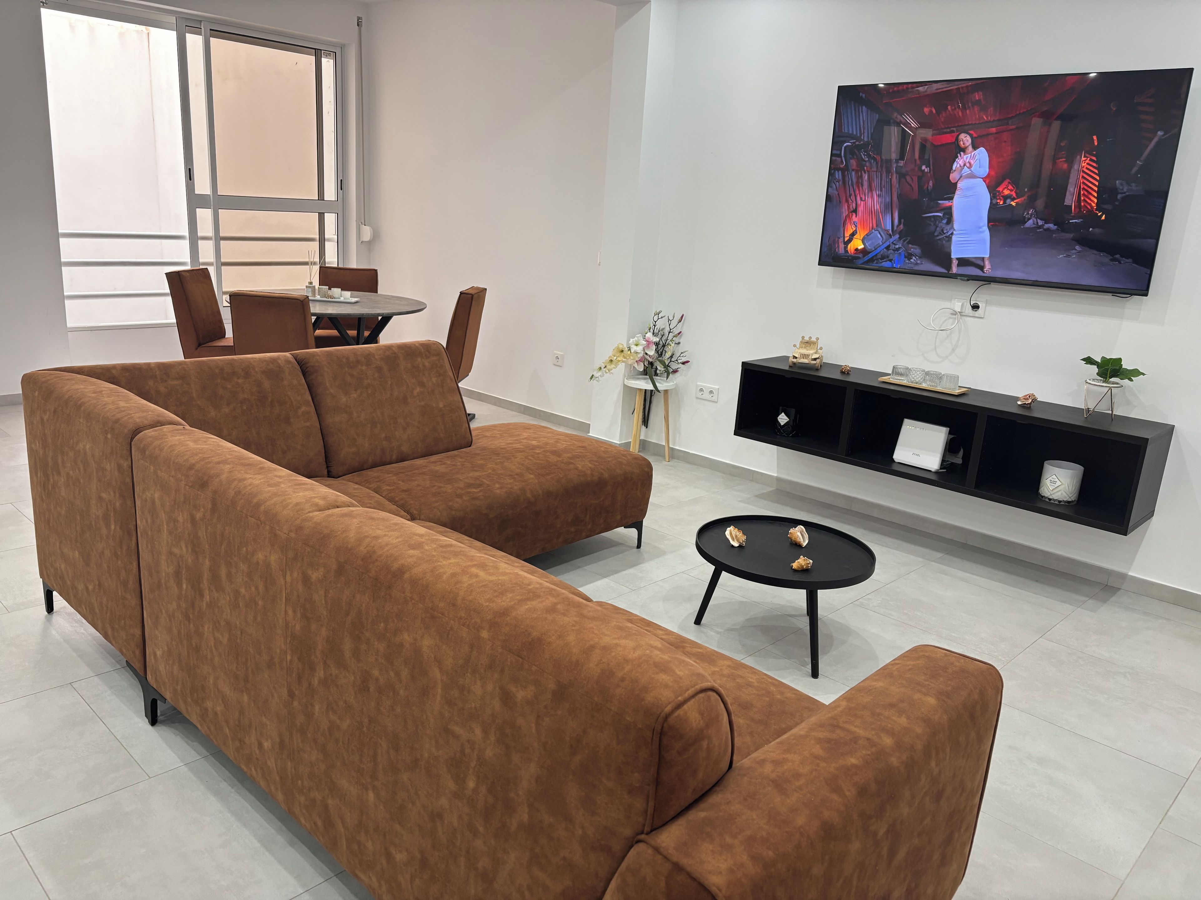 A modern living room with a brown sectional sofa, a small round table, and a flat-screen TV mounted on the wall.
