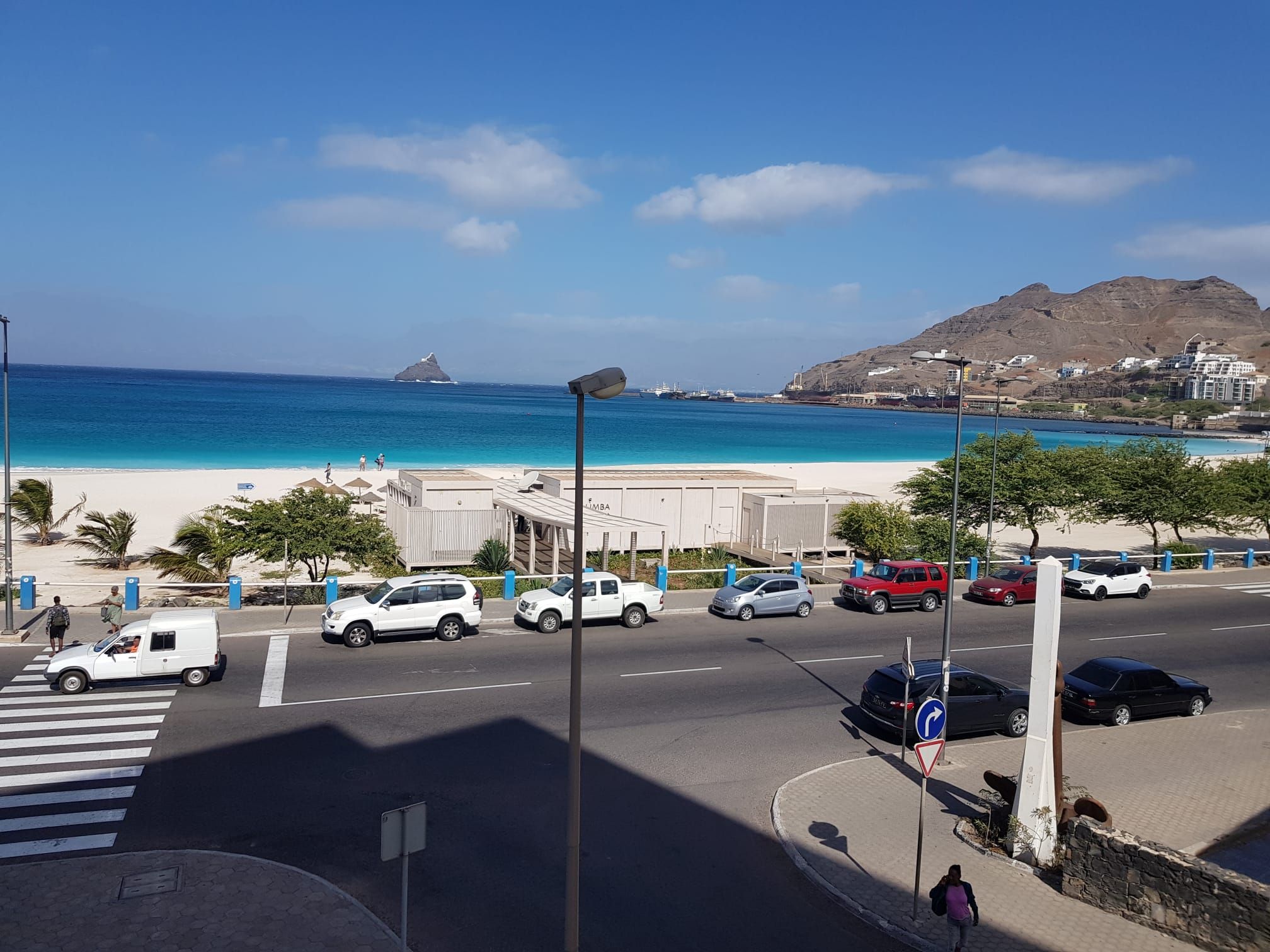 A coastal road with vehicles, a pedestrian crossing, and a beach in the background.