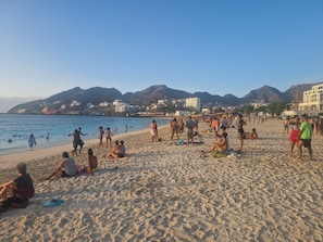 A beach with people sunbathing and swimming, buildings in the background, and mountains. - Spacious 2-Bedroom Apartment with Air Conditioning Near Laginha Beach (Mindelo)