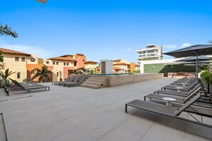 An outdoor seating area with lounge chairs and tables, a pool, and buildings in the background. - Jacana | Luxury Resort + Infinity Pool (Bucerías)