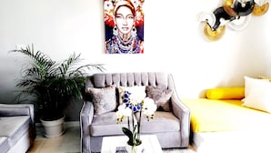 Una sala de estar con un sofá gris, una mesa de centro blanca y una cama amarilla. Hay un cuadro grande en la pared y una planta en maceta.