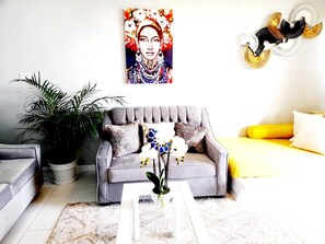 A living room with a grey sofa, a white coffee table, and a yellow bed. There is a large painting on the wall and a potted plant. - Cozy 1-bedroom apartment in fabulous Ocho Rios with AC (Ocho Rios)