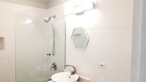 Un baño moderno con cabina de ducha de vidrio, un lavamanos blanco con encimera de mármol y un espejo hexagonal.
