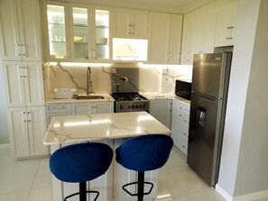 A modern kitchen with white cabinets, a stainless steel refrigerator, a gas stove, and two blue bar stools. - Cozy 1-bedroom apartment in fabulous Ocho Rios with AC (Ocho Rios)