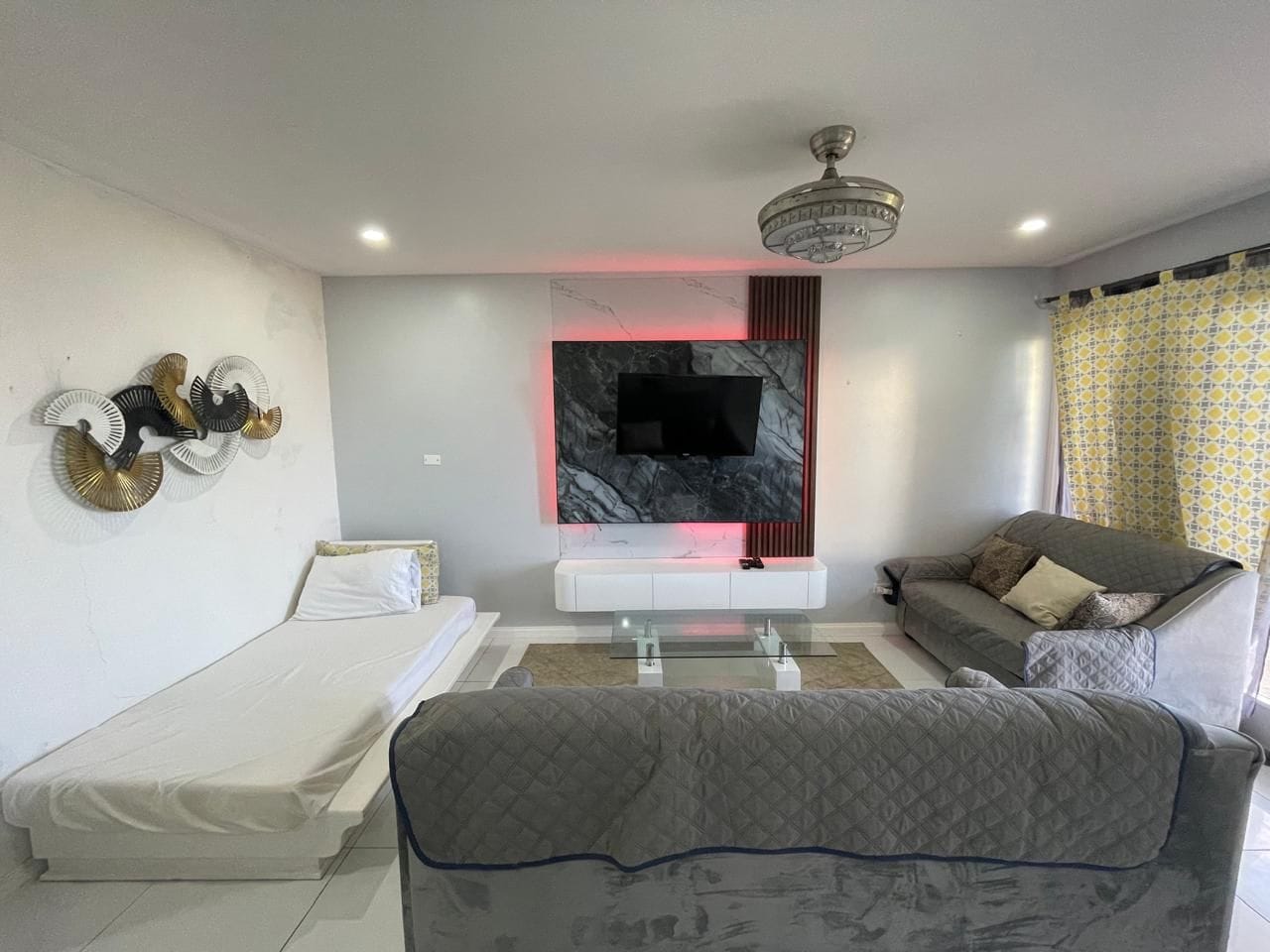 Ein modernes Wohnzimmer mit Flachbildfernseher an einer Marmorwand, einer Couch, einem Bett und einem Deckenventilator.