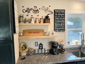 A kitchen with a coffee theme, including a sign that says 'Coffee Bar' and another that lists 'Friend Rules.' There are shelves with coffee mugs and a vintage coffee grinder.