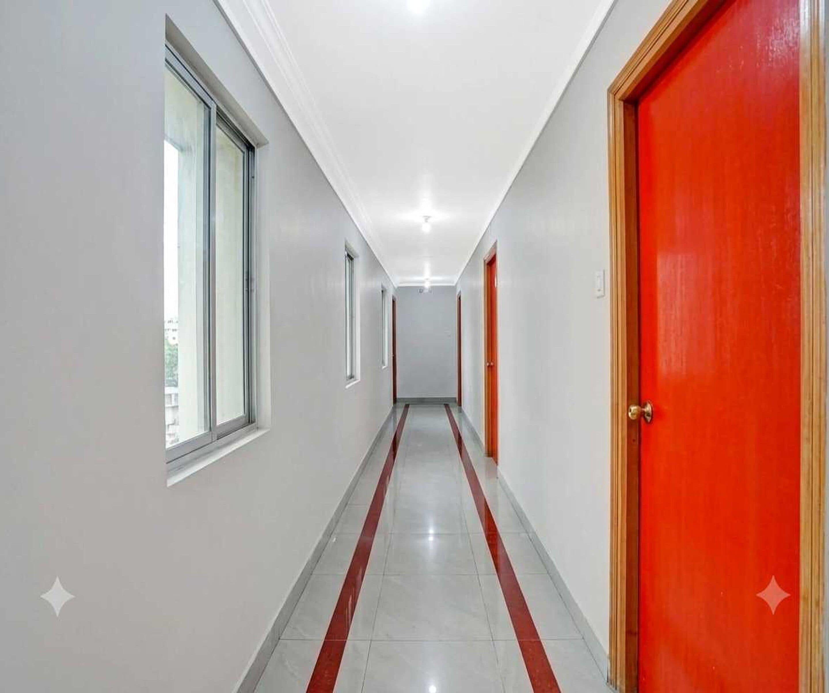 A corridor with a red door on the right, a window on the left, and a tiled floor.
