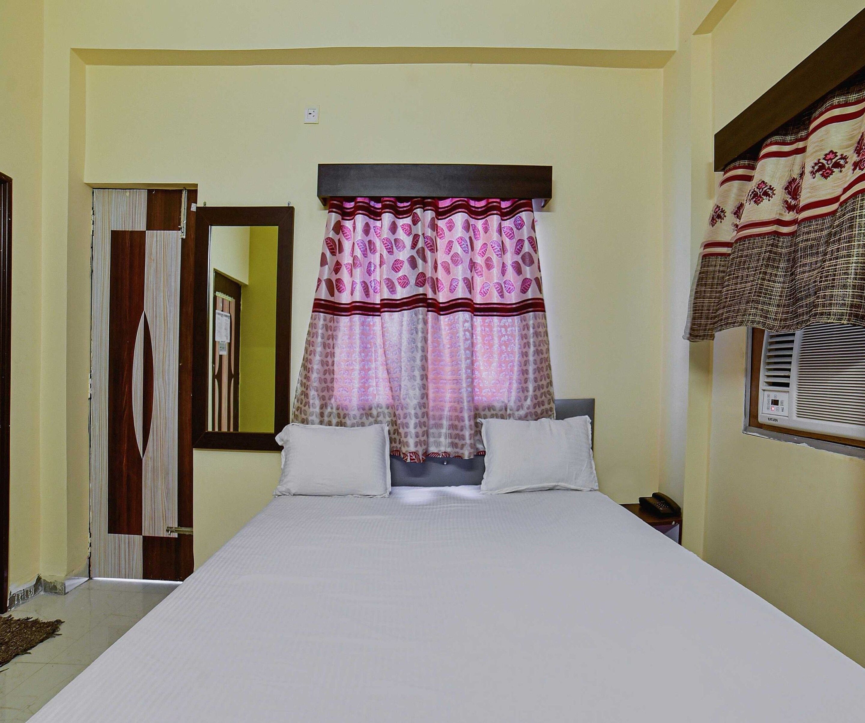 A hotel room with a bed, a telephone, a window with a curtain, and a door with a mirror.
